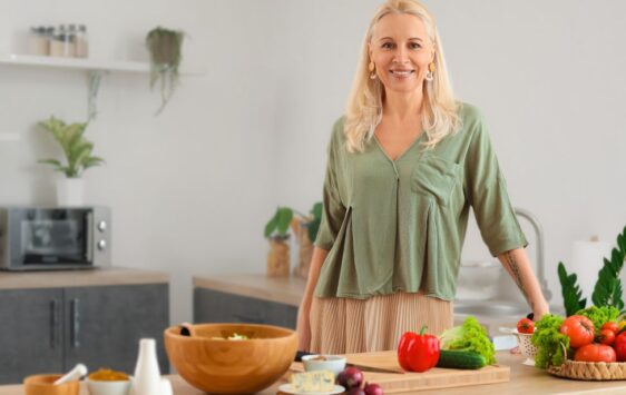 Frau in den Wechseljahren steht in der Küche hinter gesunden Lebensmitteln und lächelt dank ihrer Ernährungs- und Trainingsanpassung.