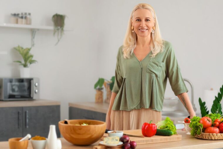 Frau in den Wechseljahren steht in der Küche hinter gesunden Lebensmitteln und lächelt dank ihrer Ernährungs- und Trainingsanpassung.